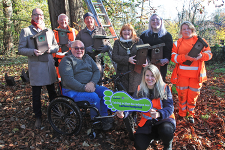 A group photo at Watnall Spinney with the habitat boxes. One person is holding a footprint shaped sign that says 'Taking action to reduce our carbon footprint'. The group includes The Mayor of Broxtowe, Councillor with Councillor Peter Bales, Ward Councillor for Watnall and Nuthall West and members of the Council’s Environment Team, along with John Parker, Chair for Nottinghamshire Bat Group and John Revill, Manager of the Nest Box Group.